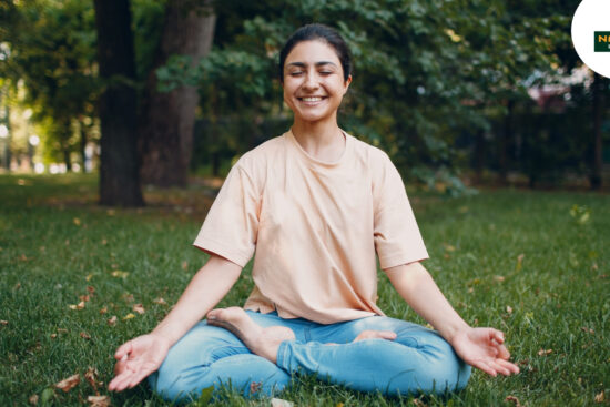 A woman peacefully meditates in a grassy field, embodying natural fitness.