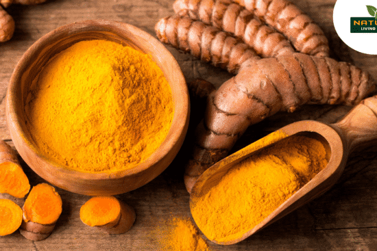 A bowl of bright yellow turmeric powder beside whole turmeric roots and a wooden scoop on a rustic wooden surface.